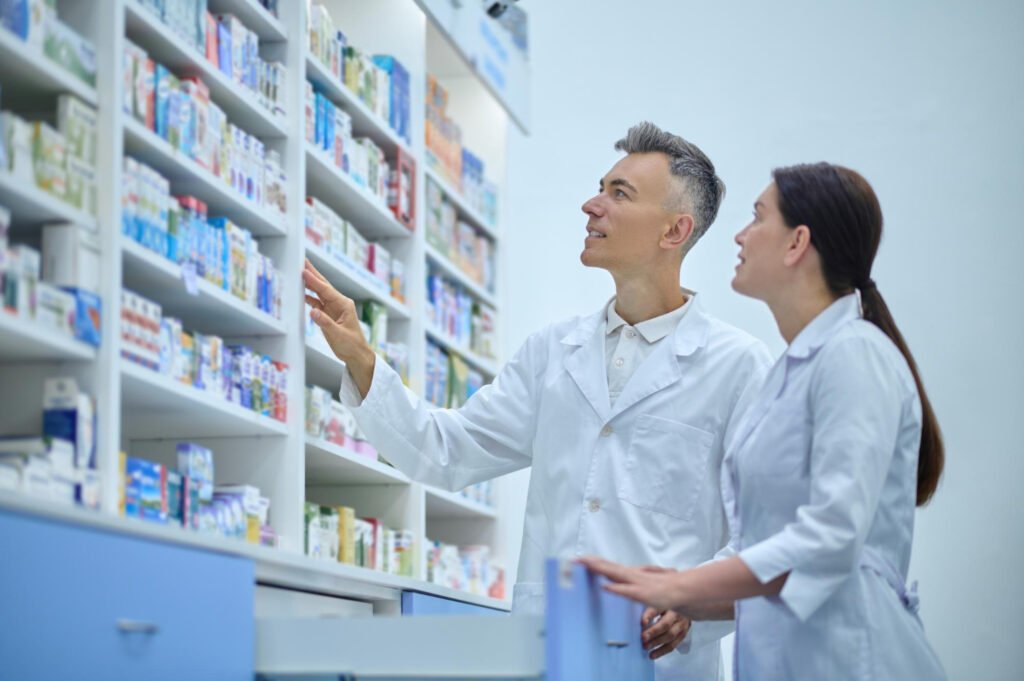 Two pharmacists in white coats discuss products in front of neatly organised pharmacy shelves in a professional setting.
